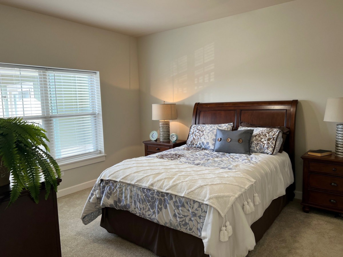A cozy bedroom featuring a neatly made bed, bedside lamp, and window with blinds.