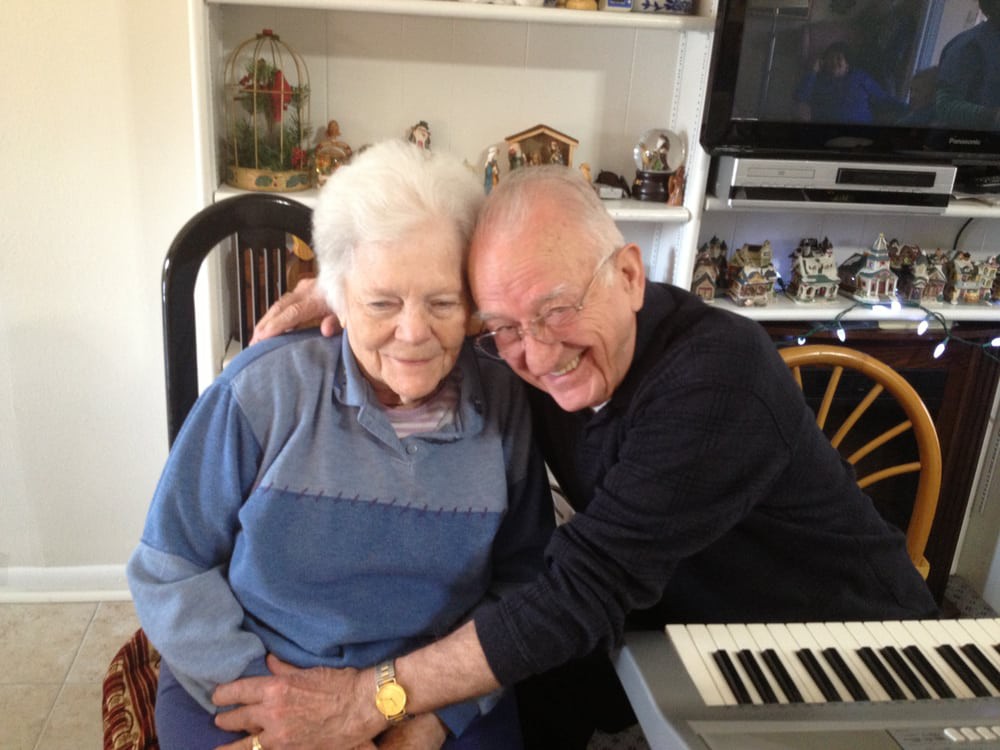 An elderly couple enjoying a warm embrace and smiling, seated next to a keyboard in a cozy home setting.