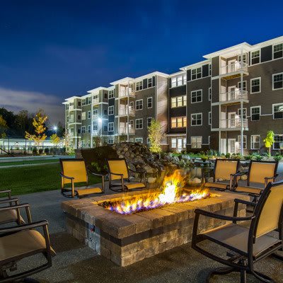 A modern apartment complex with outdoor seating around a lit fire pit during the evening.