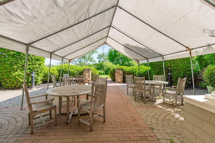 An outdoor seating area with wooden furniture under a large white tent, surrounded by greenery.
