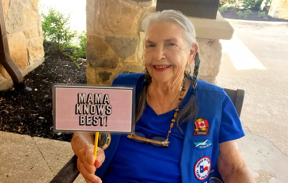An elderly woman smiling while holding a sign that says 'Mama Knows Best.'