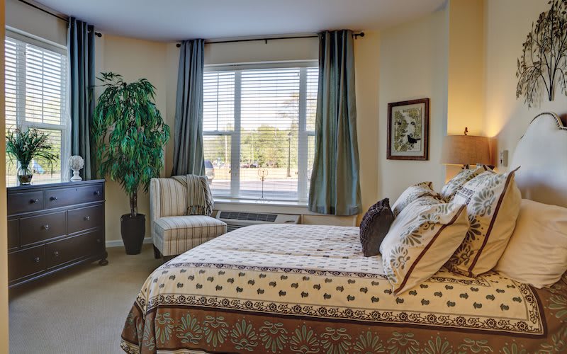 A cozy bedroom with elegant decor, featuring a large bed, upholstered chair, potted plant, and a window with curtains.