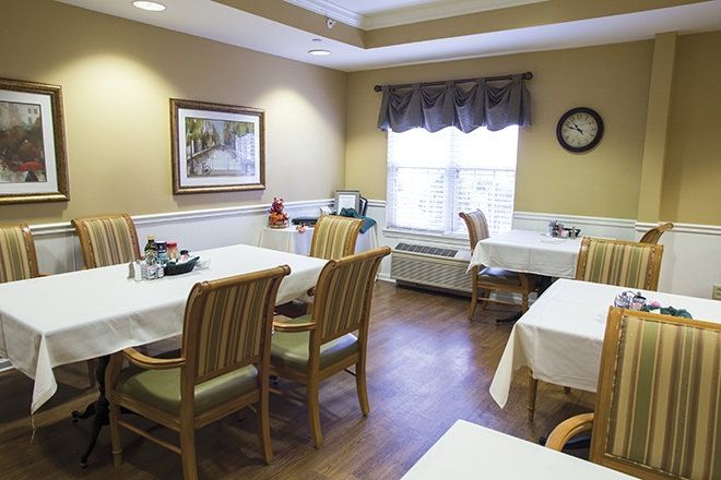 A cozy dining room with neatly arranged tables and chairs, featuring tablecloths, a large window, and wall decor.