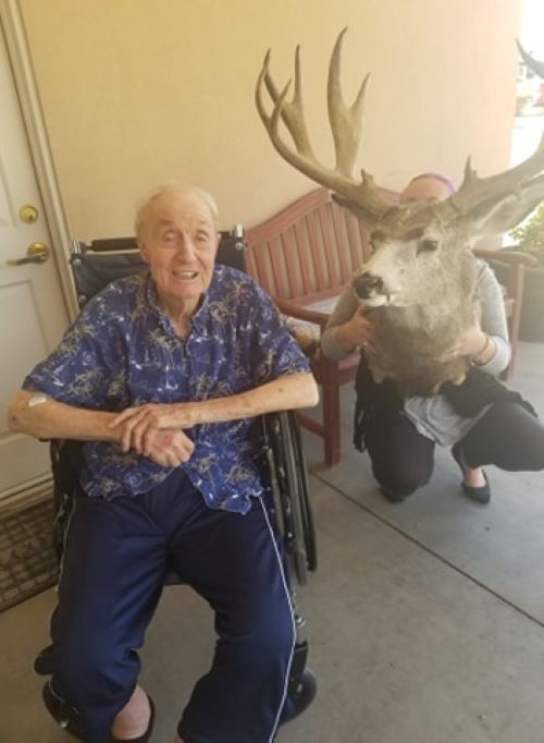 An elderly man smiling while sitting in a wheelchair indoors next to a mounted deer head.