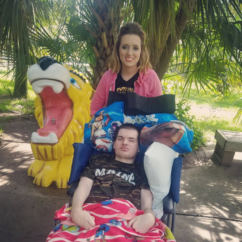 A woman stands behind a man in a wheelchair, with a colorful animal sculpture nearby in an outdoor setting.