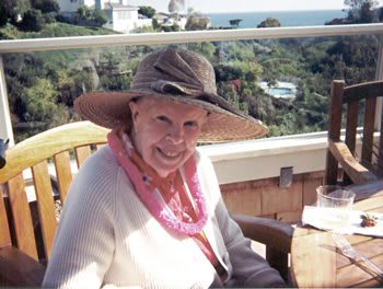 A person wearing a wide-brimmed hat is smiling while seated outdoors with a scenic view in the background.