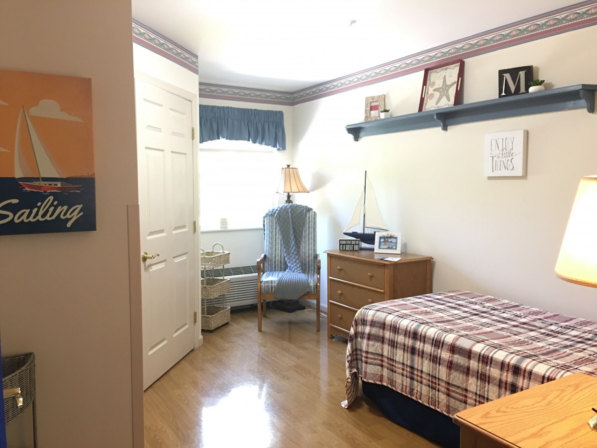 A cozy bedroom with a plaid-covered bed, wooden dresser, and a chair by the window.