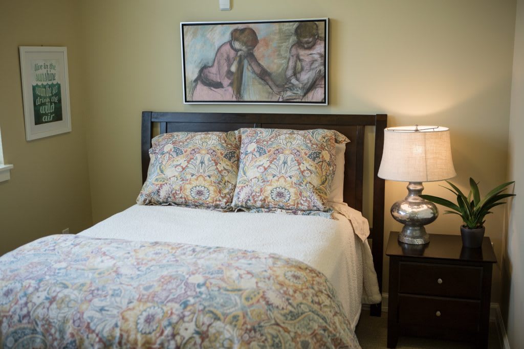 A cozy bedroom with a neatly made bed, decorative pillows and duvet, a bedside lamp, and a painting on the wall.