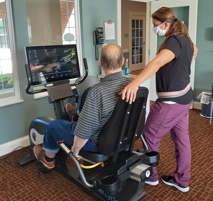 A healthcare professional assists a person on a stationary bicycle during a rehabilitation session.