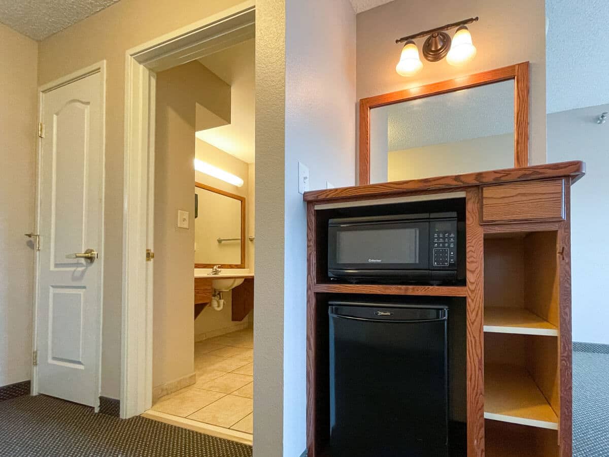 A hotel room with a view of the bathroom entrance, featuring a small kitchenette area with a microwave and refrigerator.