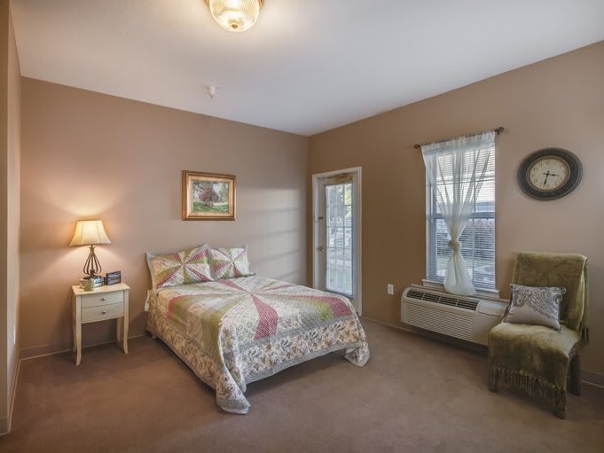 A cozy bedroom with a bed, nightstand, lamp, and armchair, featuring warm brown walls and natural light from the windows.