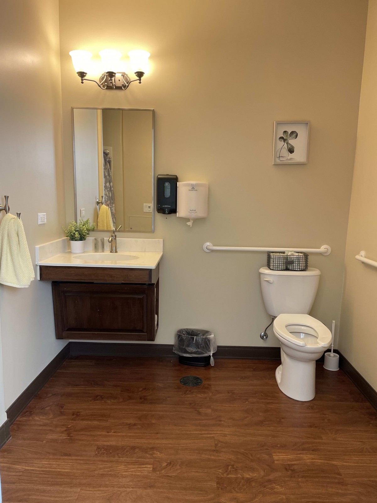 A clean bathroom with a wall-mounted sink and mirror, featuring a toilet, towels, hand sanitizer, and wall art.