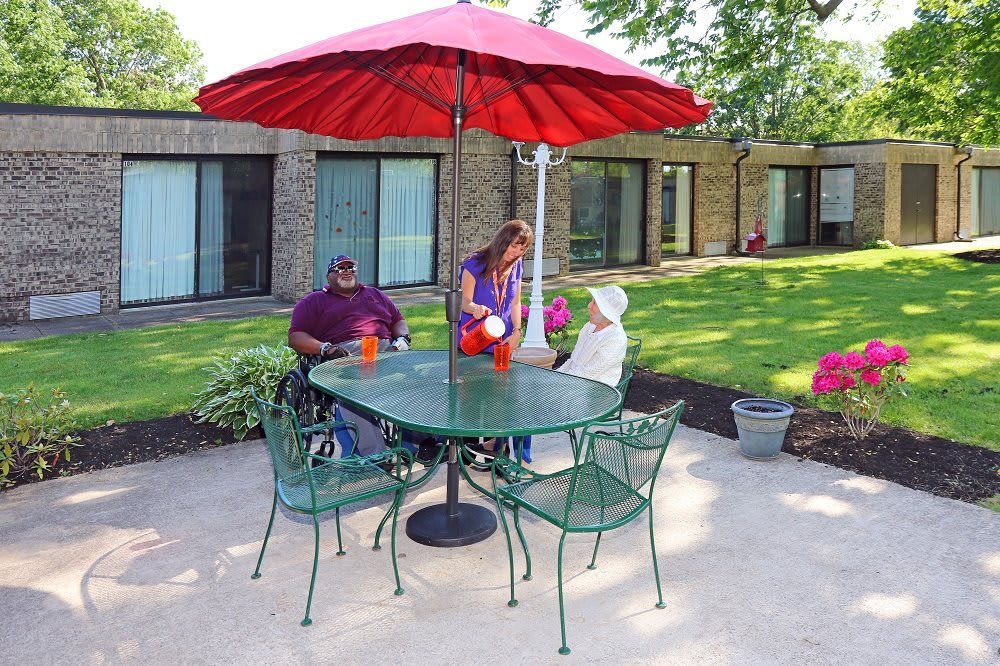 Three people sitting at a patio table under a red umbrella in a garden setting.