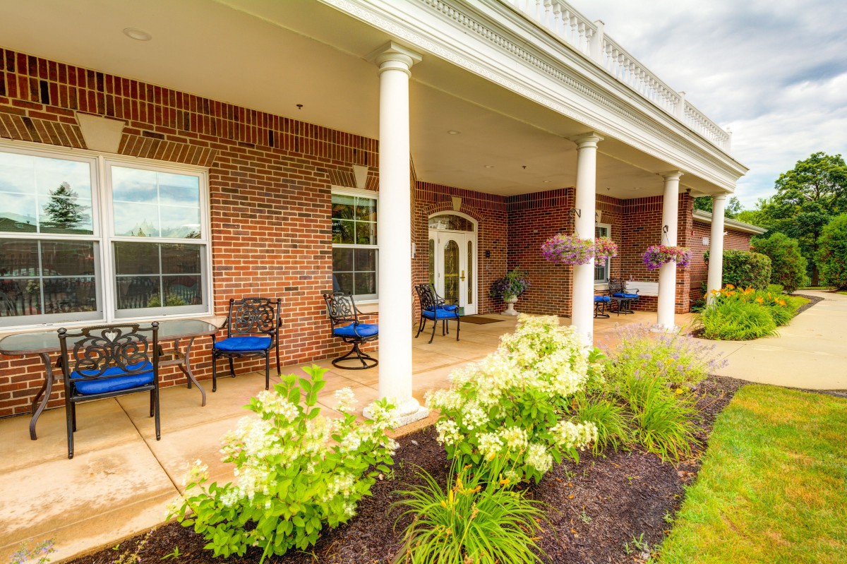 A charming brick building with a porch featuring white columns, neatly arranged chairs, and vibrant landscaping.