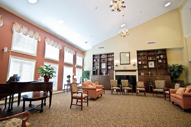 A spacious living room with a library and seating area, featuring large windows, bookshelves, a chandelier, and carpeted flooring.