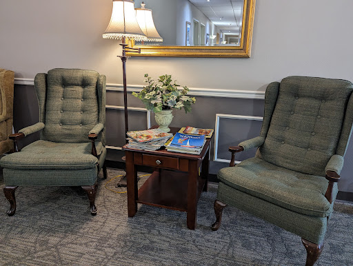 A cozy living room setting featuring two green armchairs, a wooden side table with magazines, and a lamp, complemented by a vase of flowers.