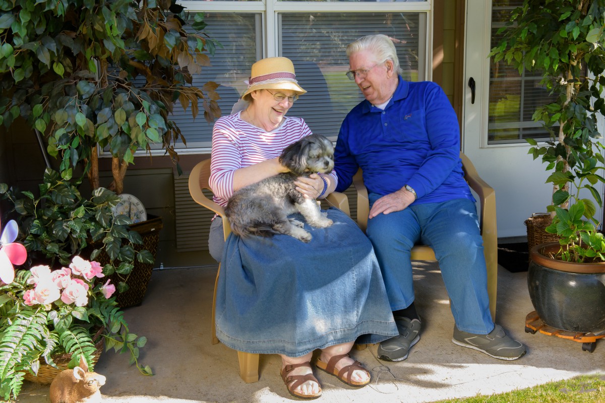 An elderly couple sitting on a porch, smiling as they enjoy time with their dog.