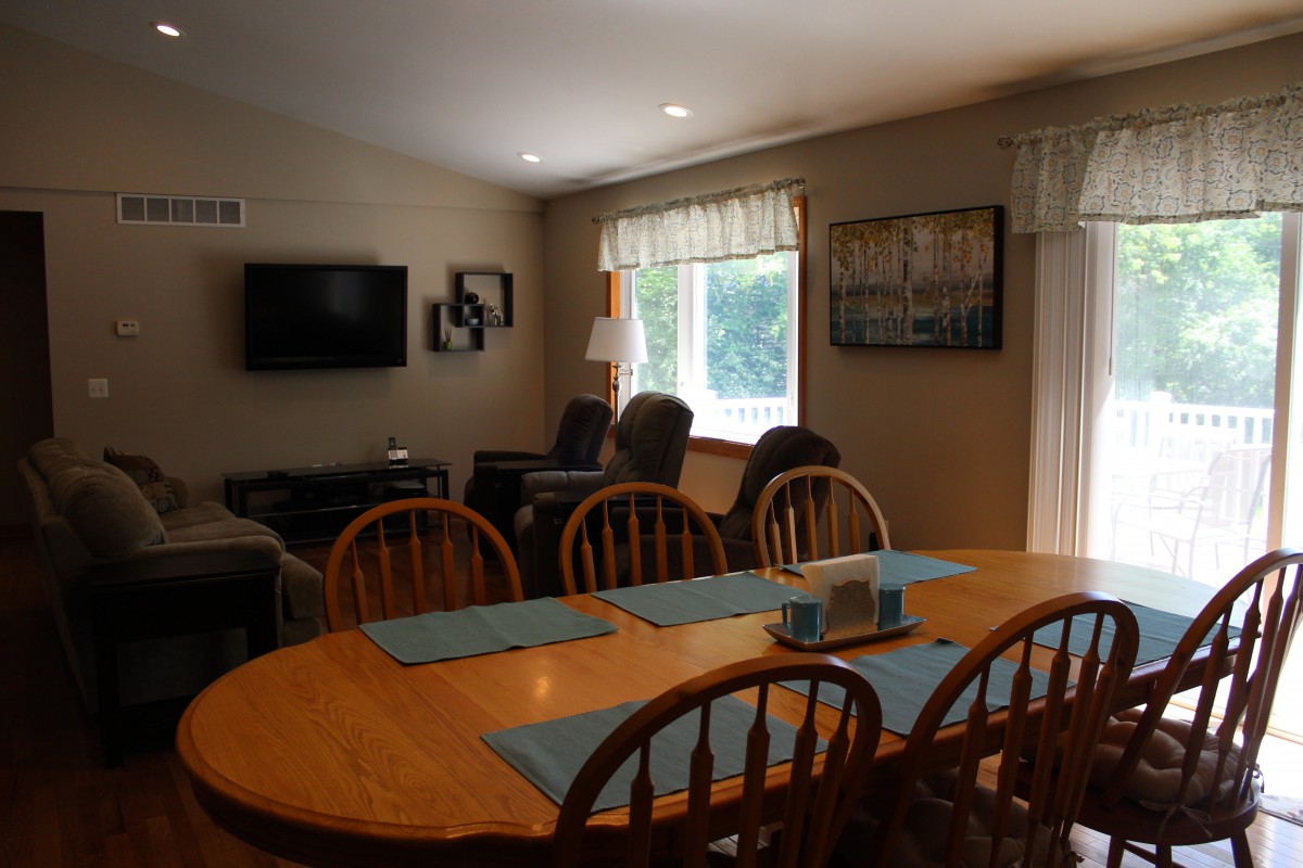 A cozy dining and living area featuring a wooden dining table with chairs, a mounted television, armchairs, and a window with curtains.