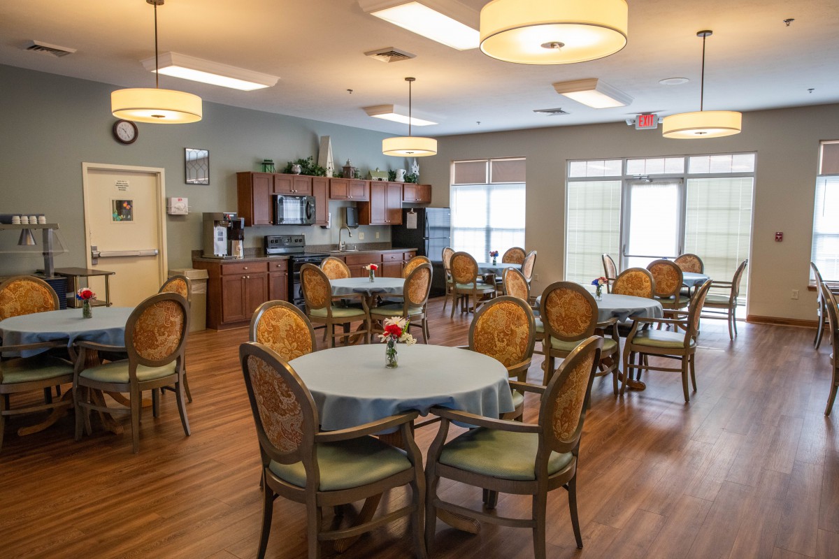 A spacious dining room with multiple tables and chairs, featuring modern lighting and a small kitchen area.