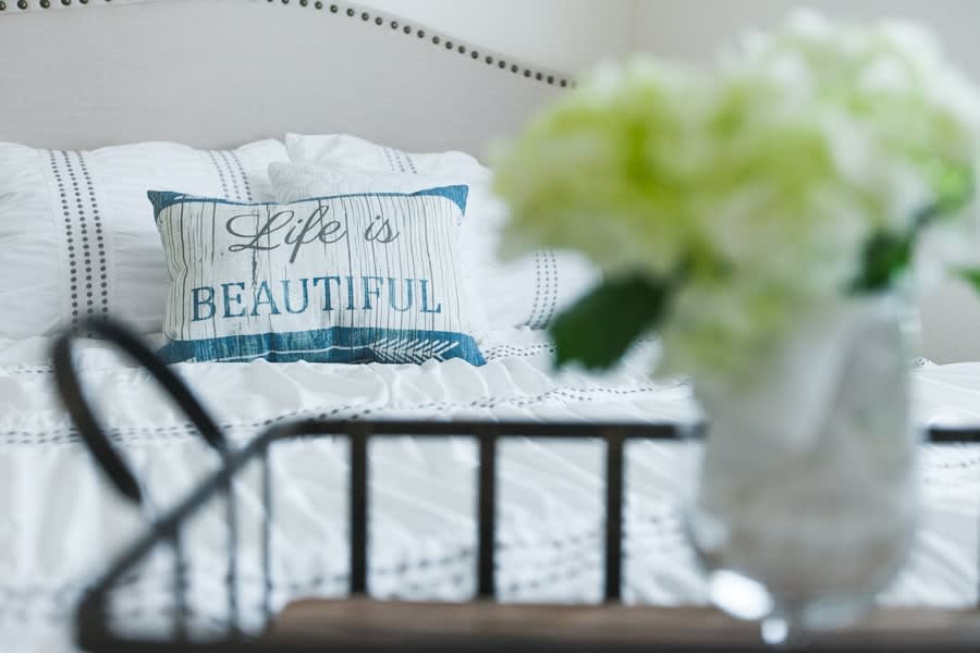 A cozy bedroom with a decorative pillow that reads 'Life is Beautiful' and a vase of flowers in the foreground.