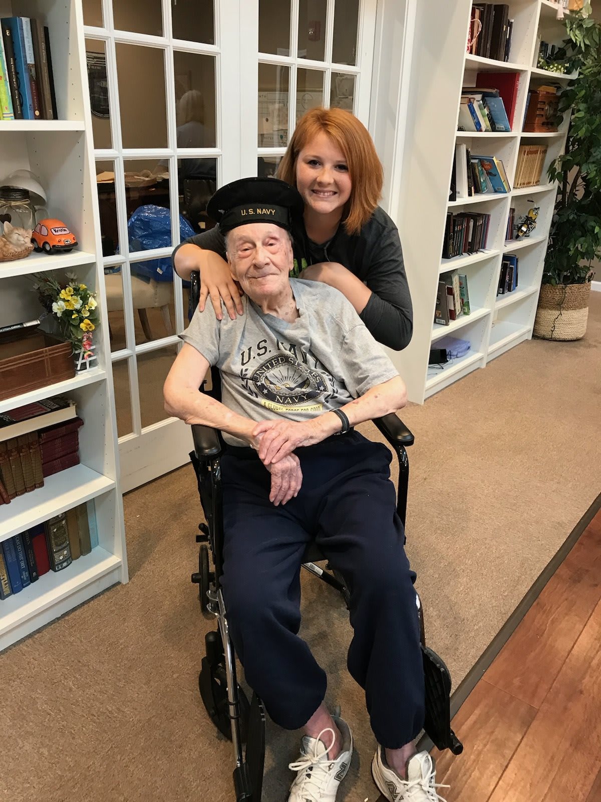 A woman smiling and posing with an elderly person seated in a wheelchair, set in a cozy library environment.