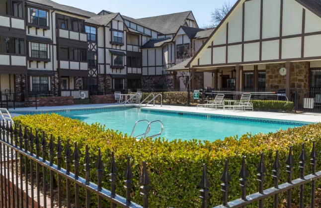 A view of an apartment complex with a swimming pool, surrounded by hedges and a decorative fence.