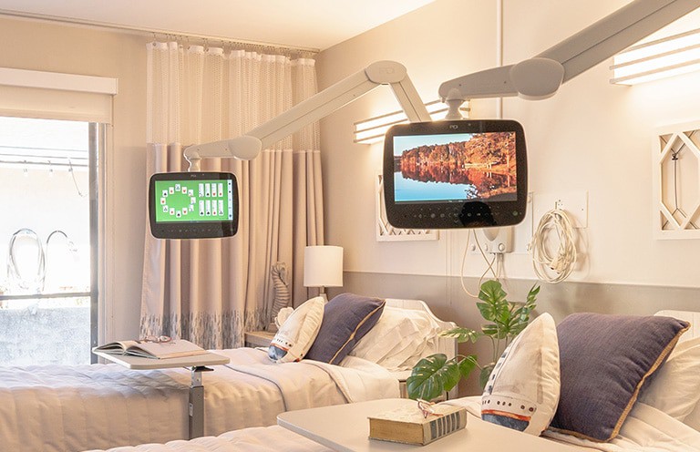 A hospital room featuring two patient beds with overhead television screens.