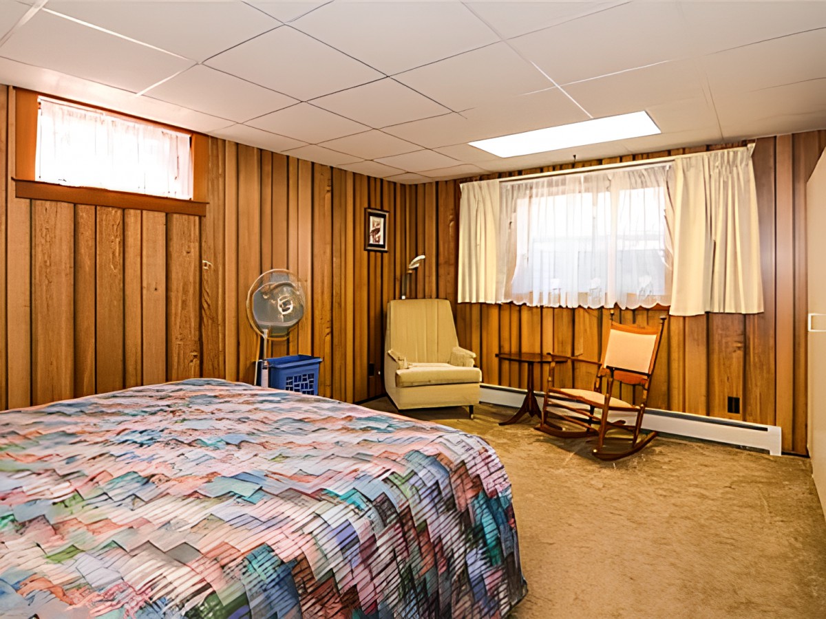 A cozy basement bedroom with wood paneling and a quilt-covered bed, featuring an armchair, rocking chair, and standing fan.