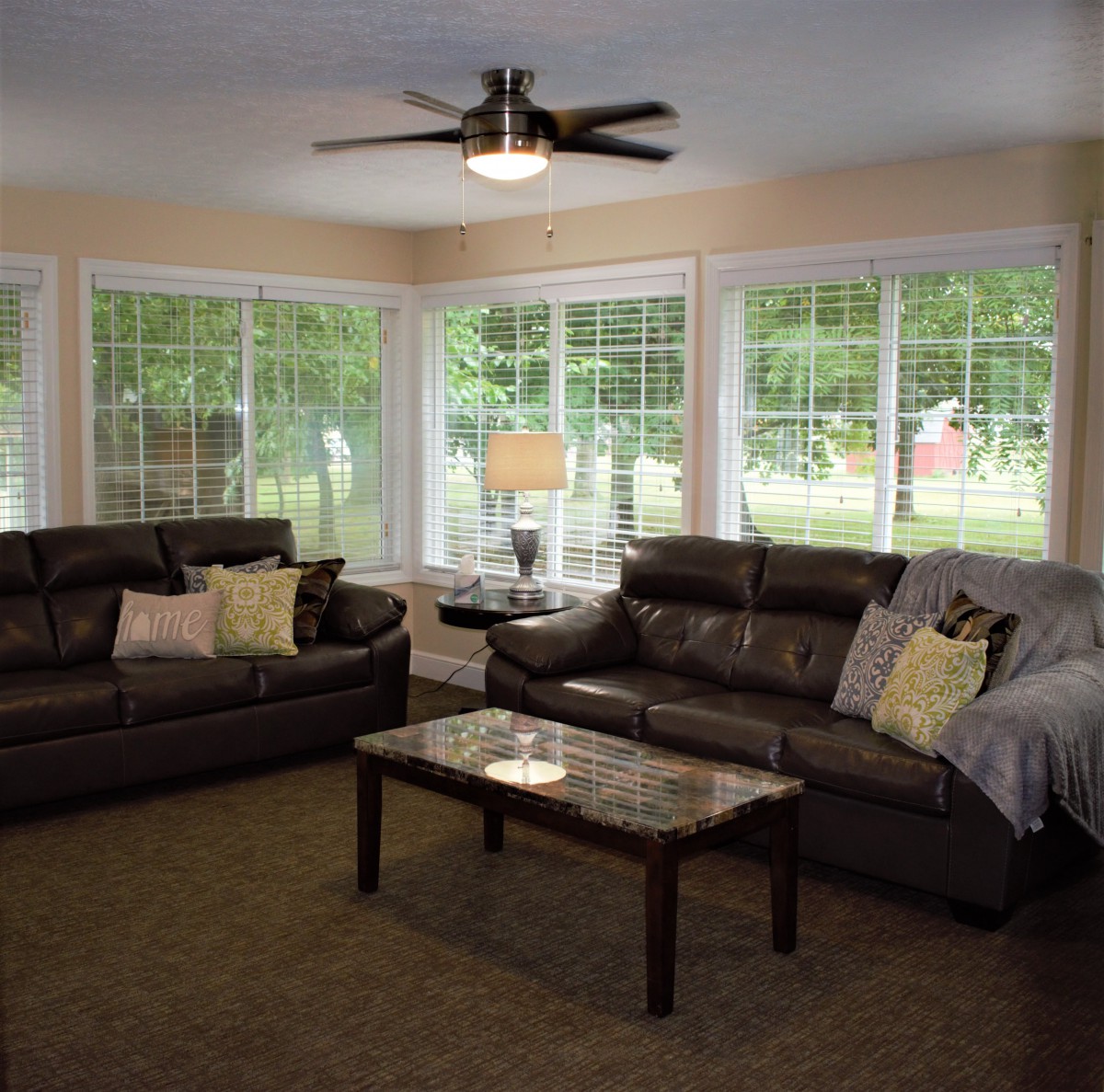 A cozy living room with large windows, two leather sofas, and a central coffee table under a ceiling fan.
