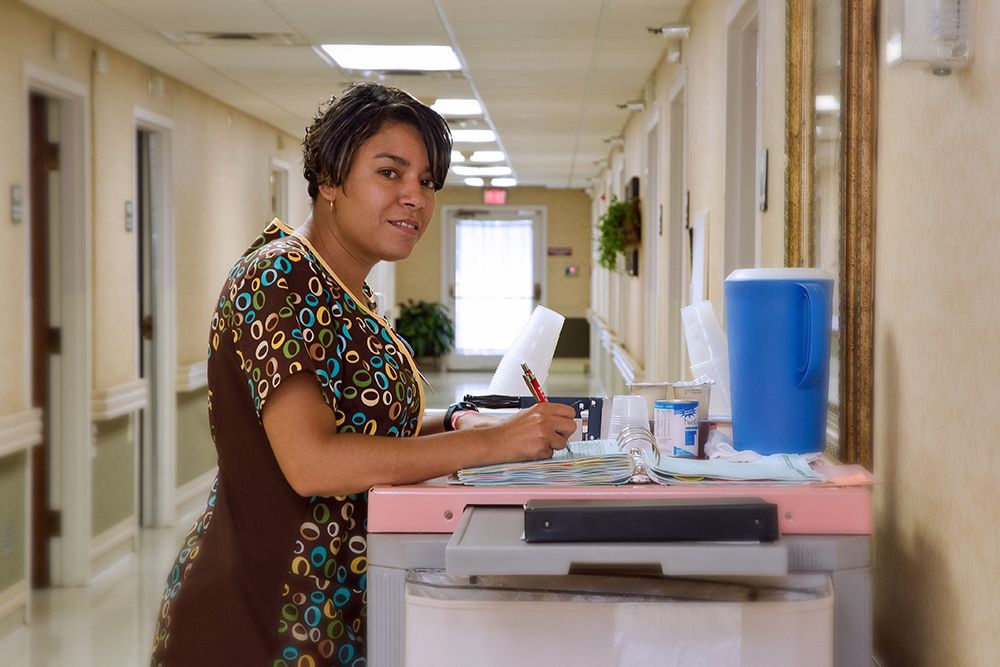 A nurse standing in a hospital corridor holding a clipboard.