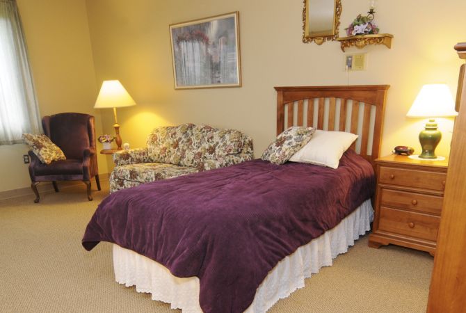 A cozy bedroom with a single bed covered in a purple blanket, accompanied by a floral sofa and a lamp.