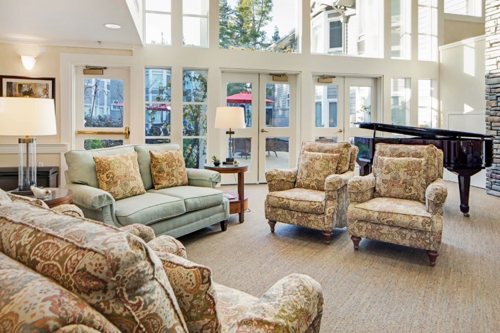 A cozy living room with plush sofas and chairs, a piano, large windows, and a lamp adding warmth to the space.