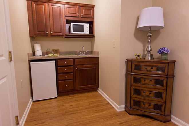 A cozy kitchenette with wooden cabinets, a microwave, mini fridge, and a decorative lamp on a drawer unit.