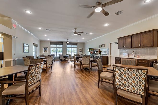 A spacious dining area with wooden floors, multiple tables and chairs, ceiling fans, and large windows.