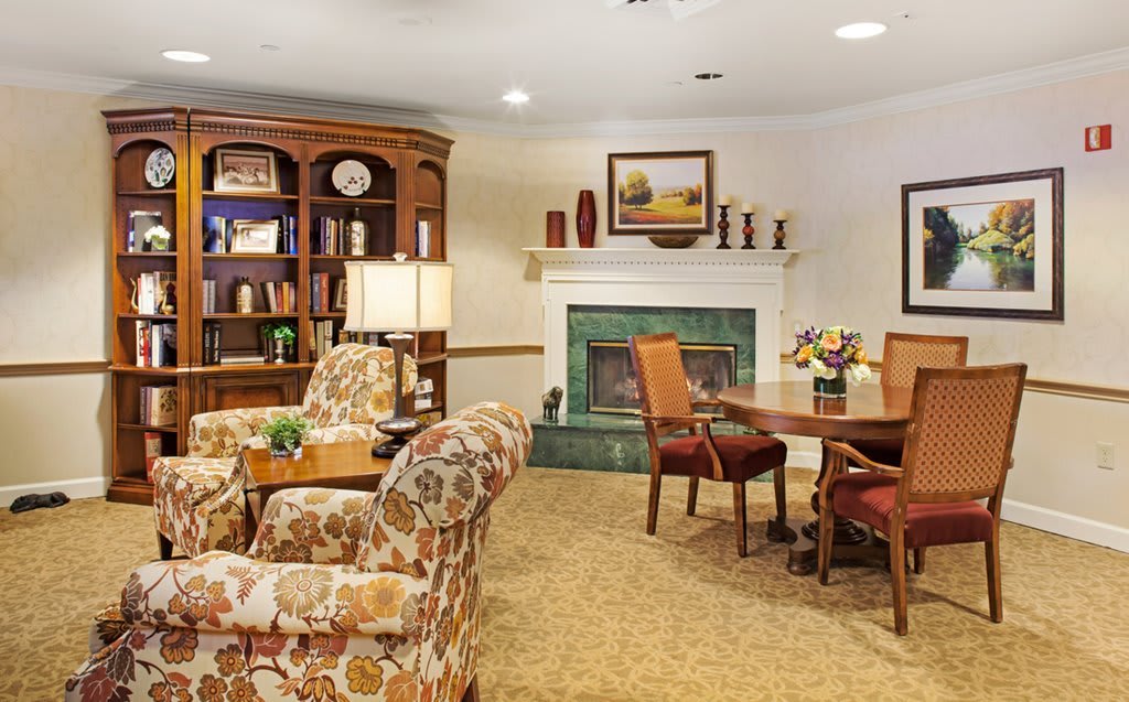 A cozy living room with a fireplace, armchairs, a bookshelf, and a table.