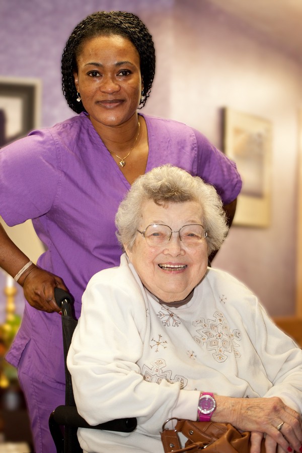 A caregiver standing behind an elderly woman sitting in a wheelchair, both smiling warmly.