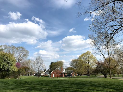 A serene view of a park with lush green grass, trees in bloom, and a charming building under a partly cloudy sky.