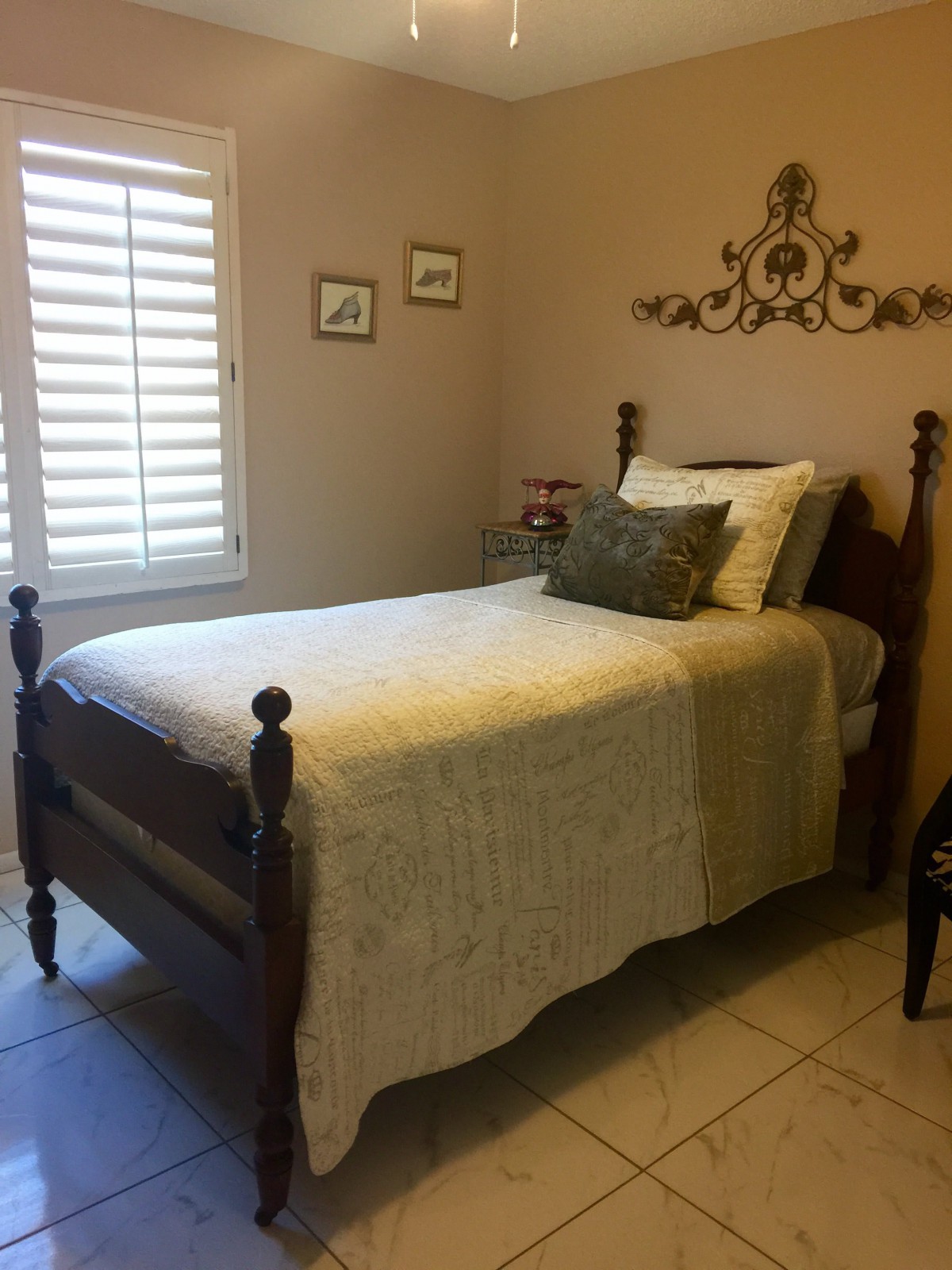 A cozy bedroom with a single traditional style bed, adorned with pillows and decorative wall art, illuminated by natural light through window shutters.