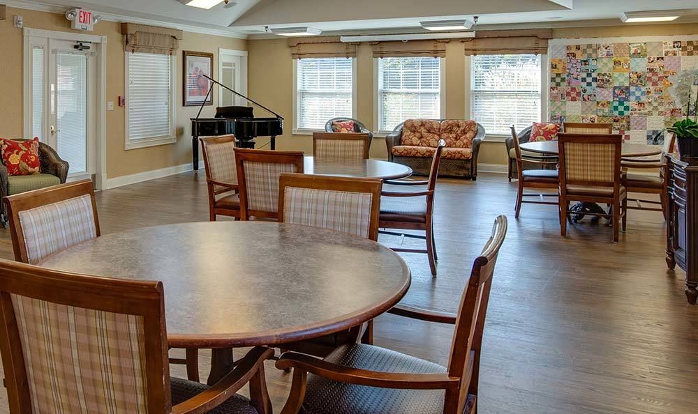 A community room with tables, chairs, and a piano near large windows.