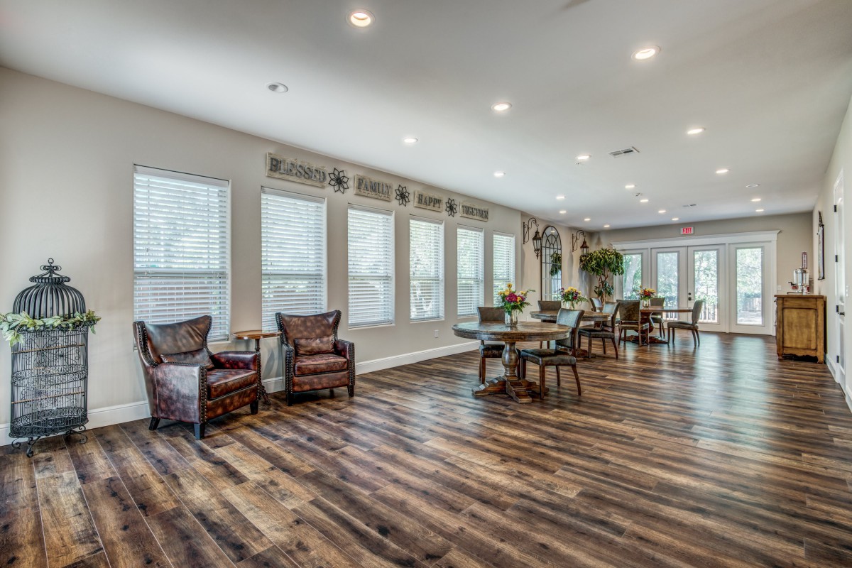 A spacious living room with wooden flooring and ample natural light shining through large windows, furnished with chairs and tables.