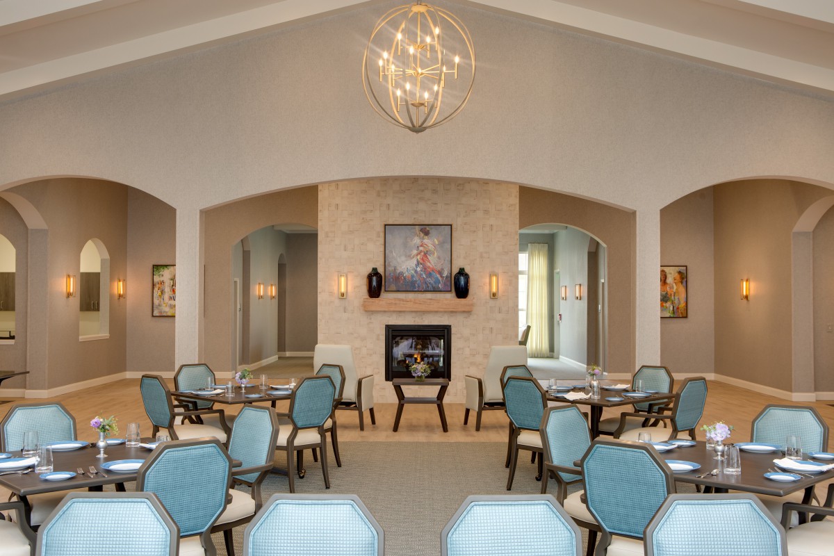 A spacious and modern dining room with elegant blue chairs, a central fireplace, and a large chandelier overhead.