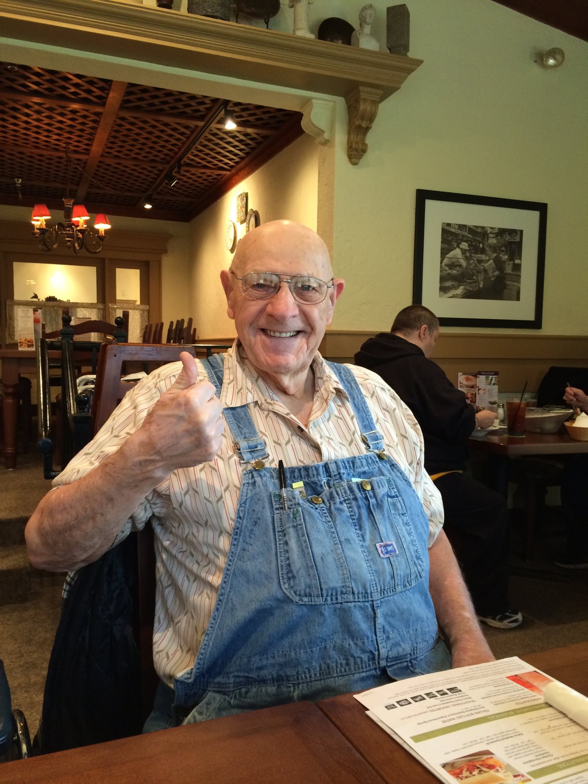 A cheerful man in overalls gives a thumbs-up at a restaurant.