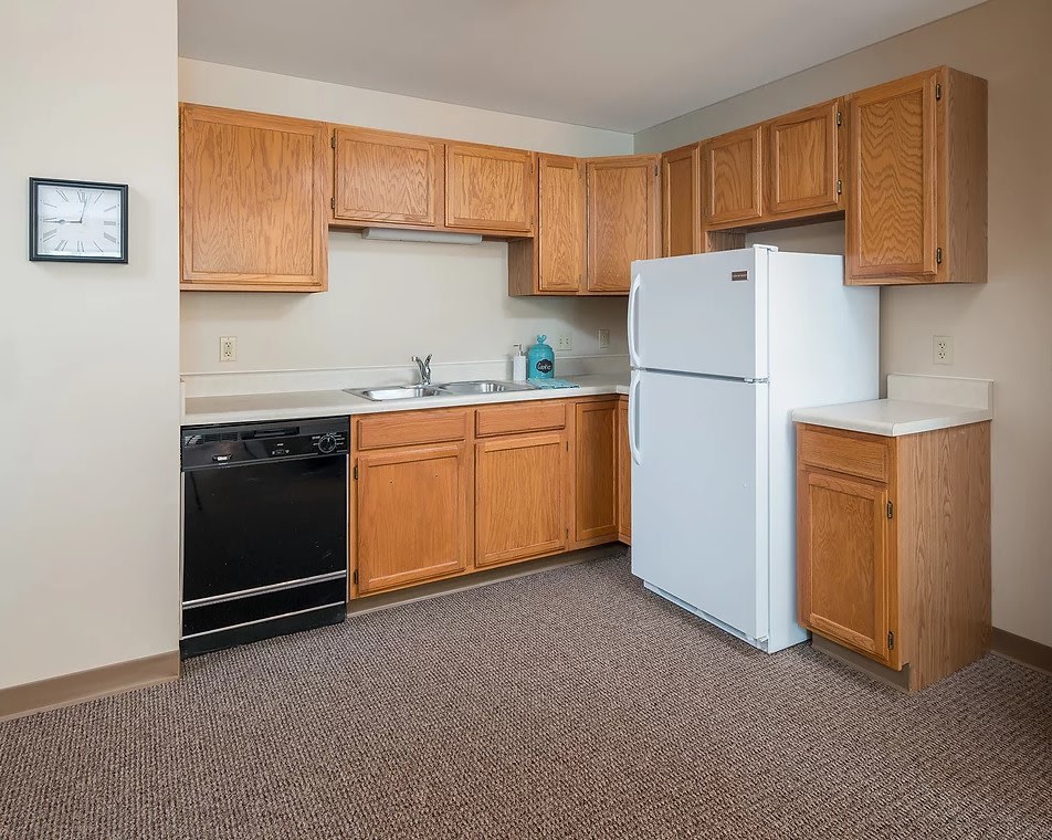 A modern apartment kitchen with wooden cabinets, a refrigerator, and carpeted flooring.
