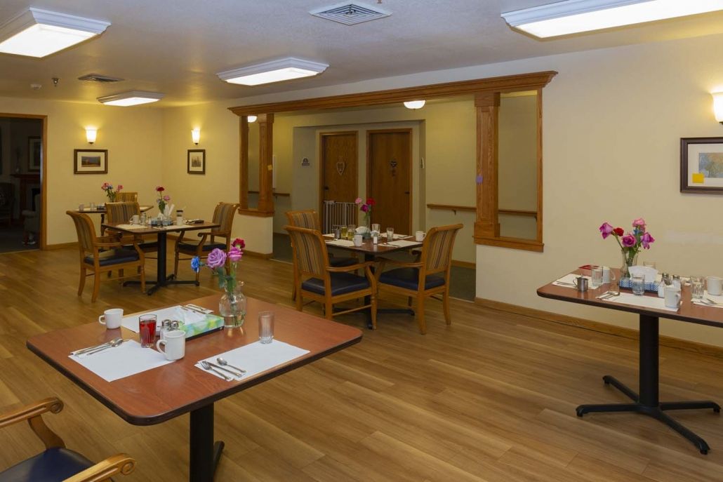 A cozy dining room with neatly arranged tables and chairs, featuring wooden floors and flower arrangements.