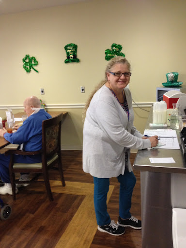 A cheerful woman assists in a senior living community, surrounded by festive green decorations.