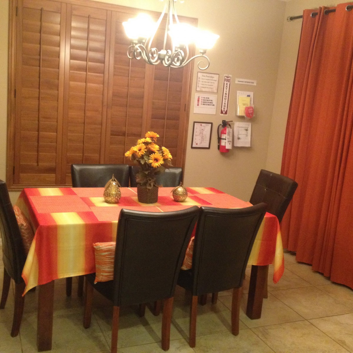 A cozy dining room featuring a table with a colorful tablecloth, surrounded by chairs and decorated with a vase of sunflowers under a chandelier.