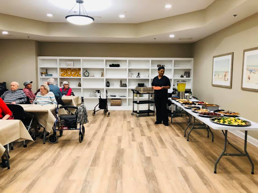 A community activity room in a senior living facility, featuring residents engaging with staff amidst a table of refreshments.
