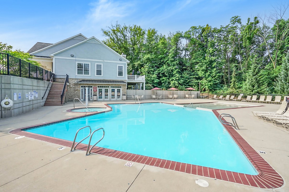 A serene outdoor swimming pool adjacent to a residential house, surrounded by lush trees and a clear blue sky.