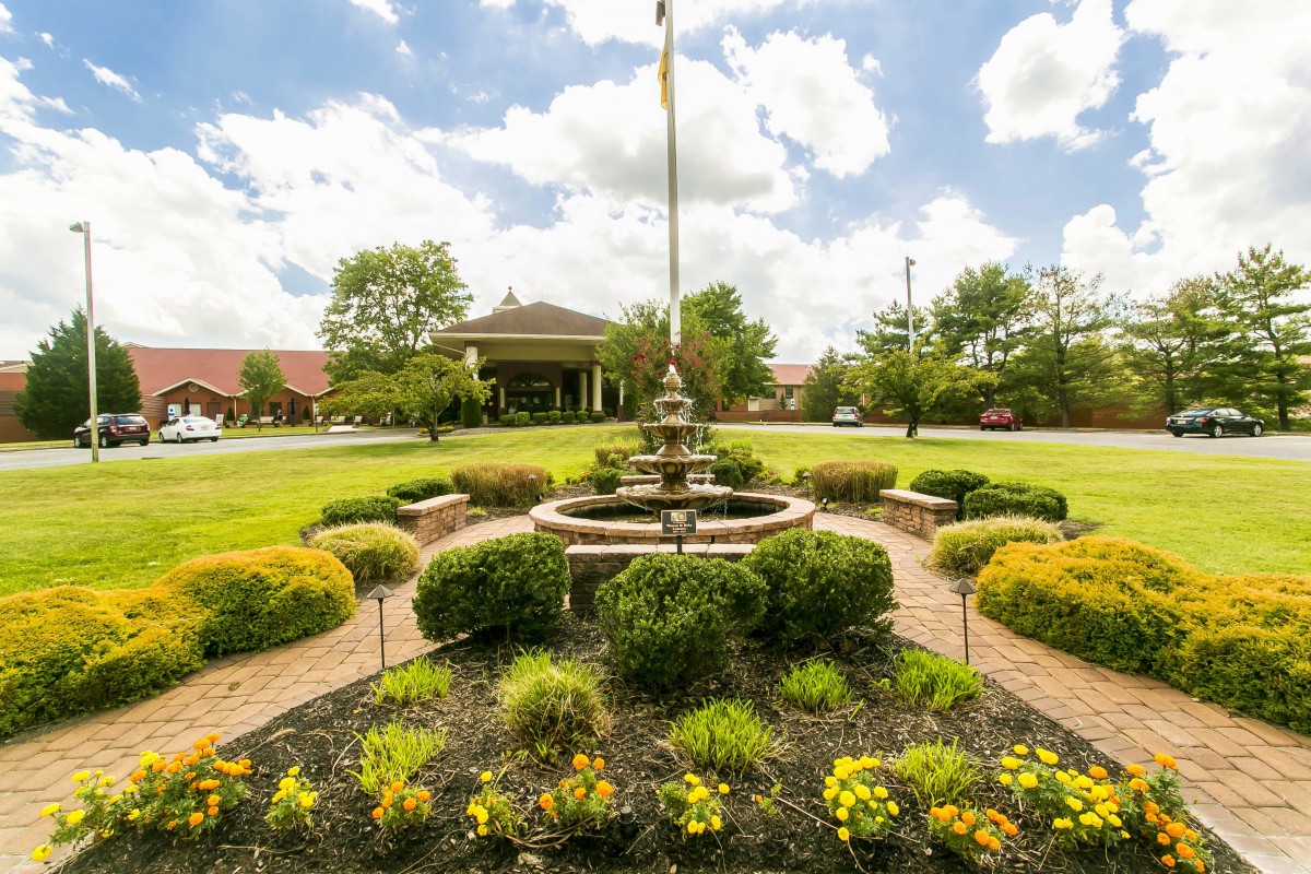 A beautifully landscaped garden with a central fountain surrounded by lush greenery and colorful flowers under a cloudy sky.
