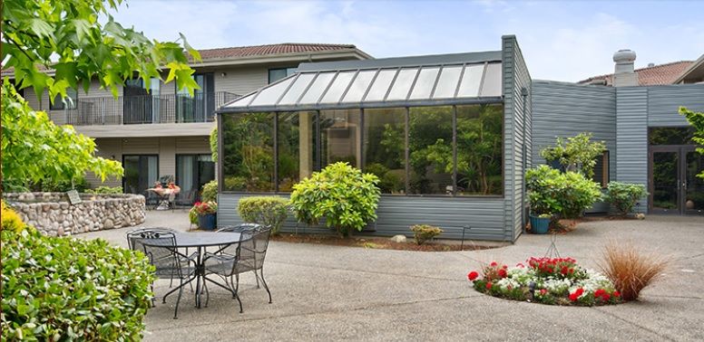 A modern conservatory with glass walls, surrounded by lush greenery and an outdoor patio area with seating.